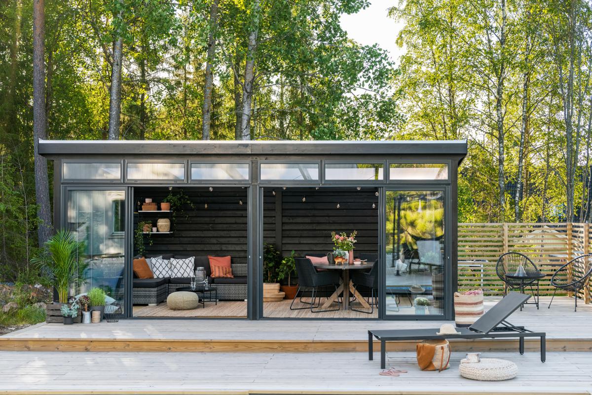 Bild på fristående uterum Önska från Skånska Byggvaror. Uterummet står på ett trädäck vid en pool med gröna träd i bakgrunden.